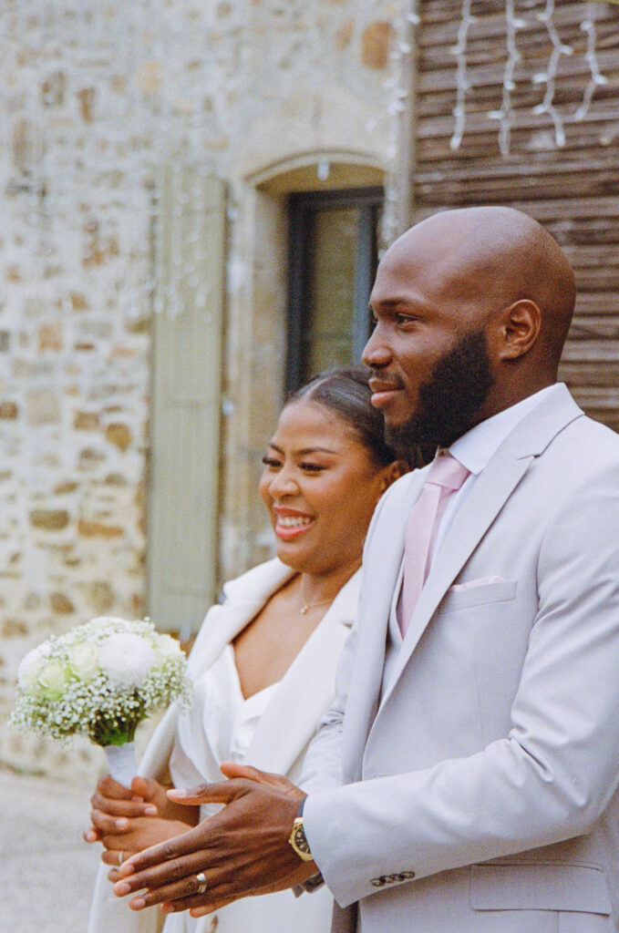 Couple de mariés en tenue blanche élégante, la mariée tenant un bouquet de fleurs blanches, capturé en noir et blanc.