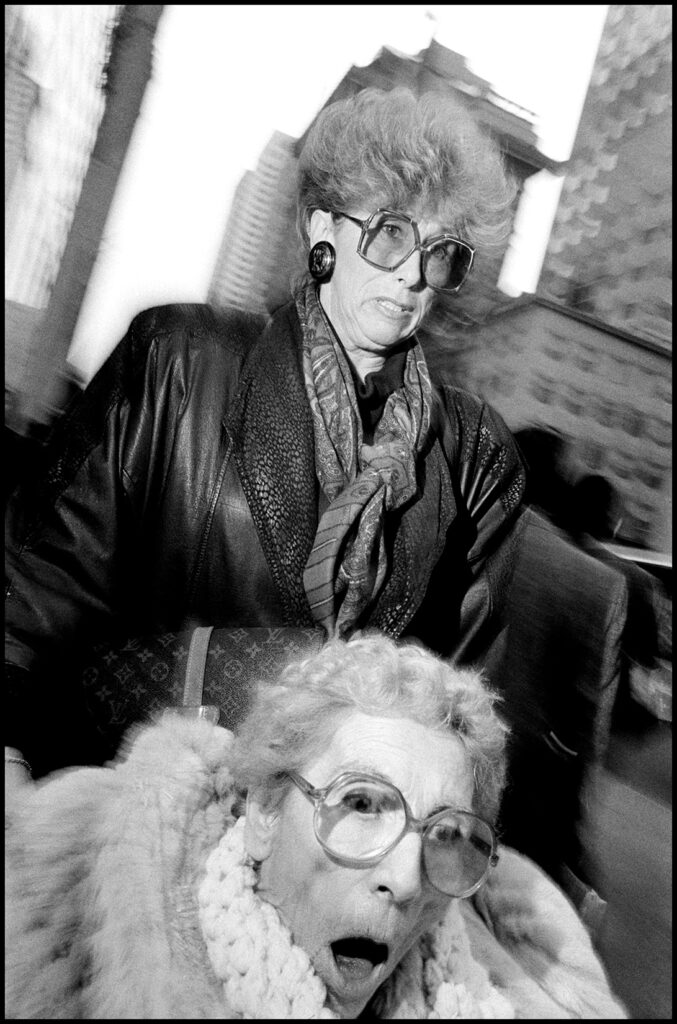 Photographie en noir et blanc de Bruce Gilden capturant deux femmes âgées dans la rue, avec des expressions marquées et des lunettes de soleil, exemple typique de sa photographie de rue crue et directe.