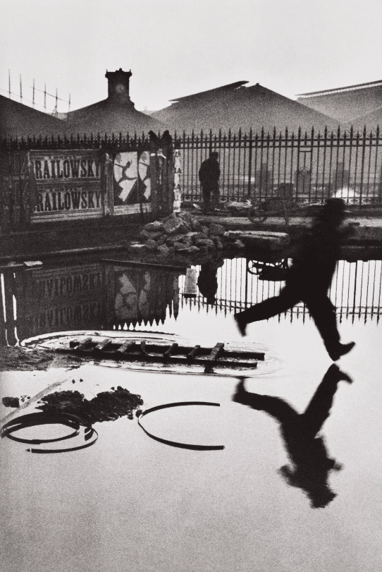 Photo en noir et blanc d'Henri Cartier-Bresson, capturant un homme sautant au-dessus d'une flaque d'eau derrière la gare Saint-Lazare à Paris en 1932.