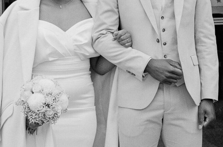 Couple de mariés en tenue blanche élégante, la mariée tenant un bouquet de fleurs blanches, capturé en noir et blanc.