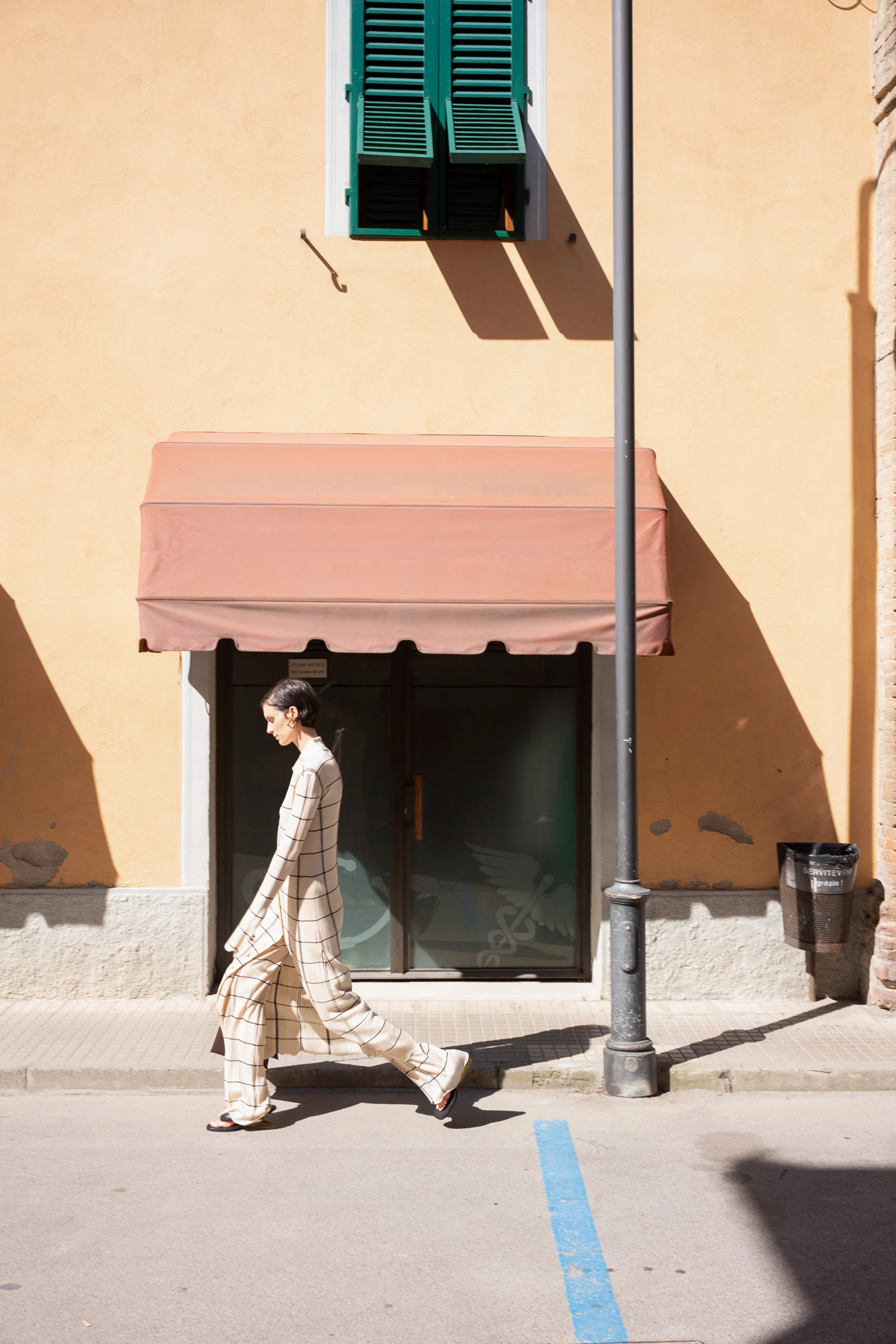 Photographie de rue par Joel Meyerowitz pour la campagne Jil Sander FW21, montrant une femme en tenue à carreaux marchant sous une lumière naturelle devant un bâtiment beige.