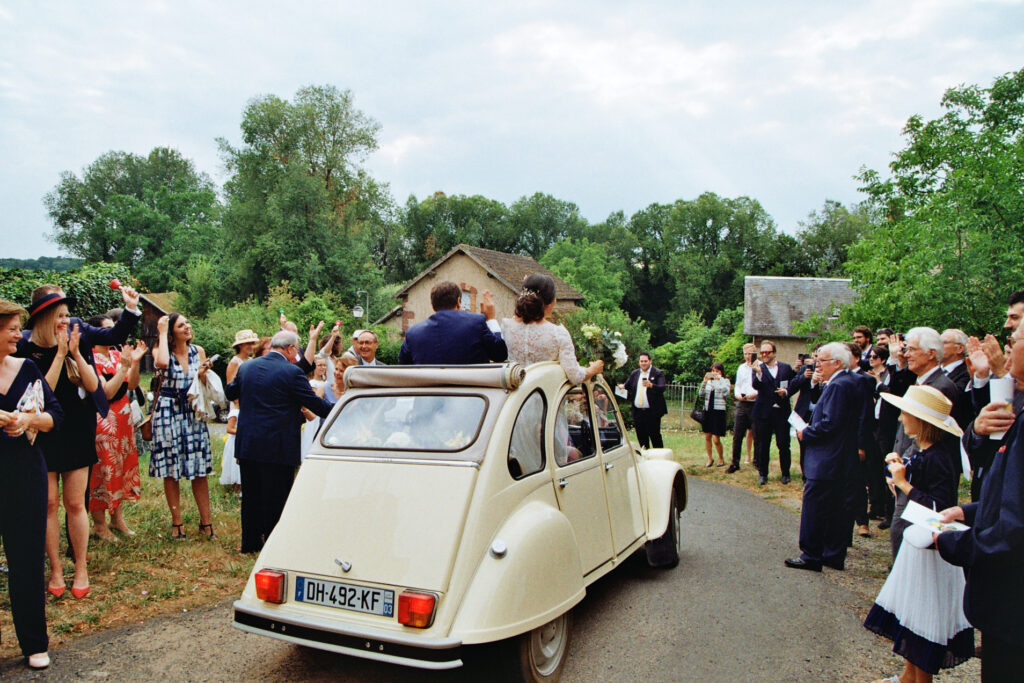 Mariés quittant leur cérémonie dans une voiture vintage, salués par leurs invités.