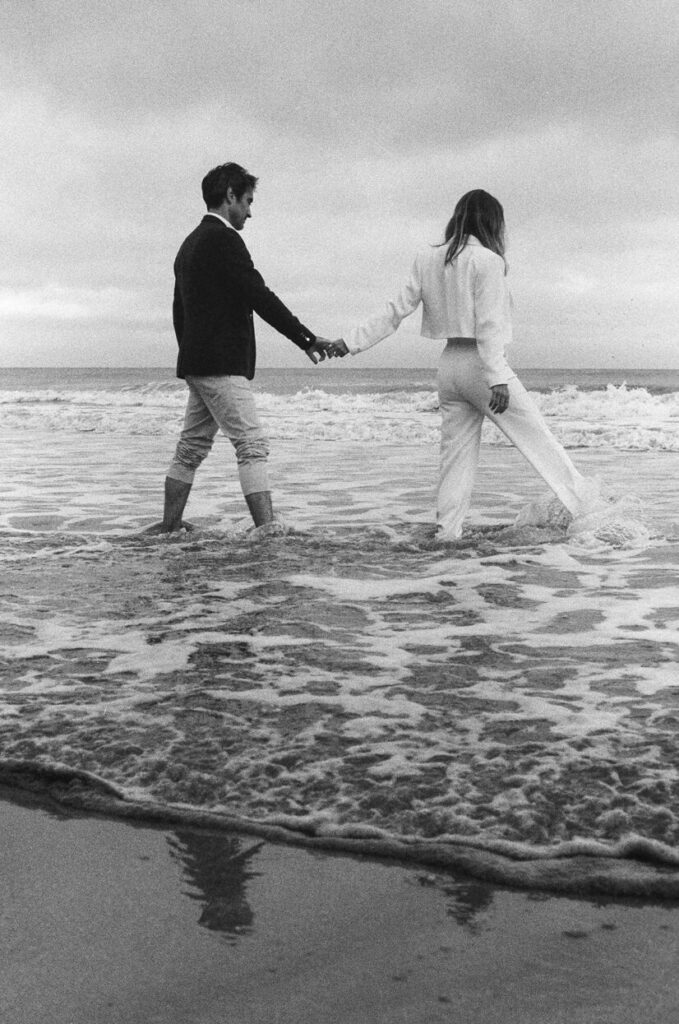 Couple marchant main dans la main dans les vagues sur une plage, capturé en noir et blanc.